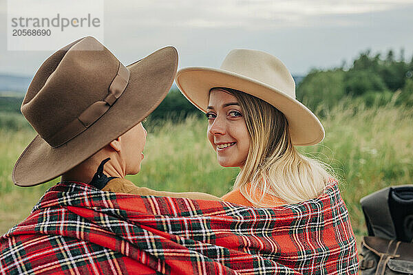 Smiling young woman wearing hat and sitting with boyfriend in plaid shawl at meadow