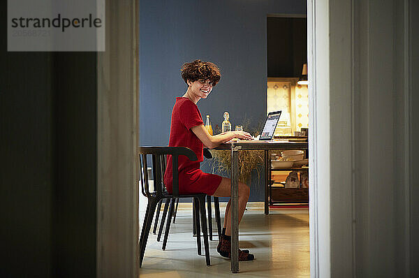 Smiling young woman sitting with laptop near table at home