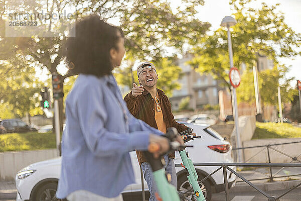 Laughing man pointing at girlfriend and standing with electric push scooter at park