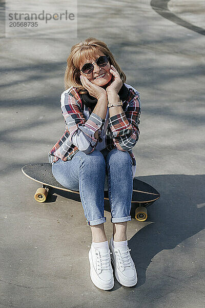 Happy senior woman sitting on skateboard at sports ramp in park