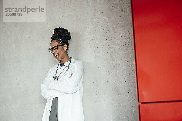 Cheerful young female doctor with arms crossed leaning on wall at hospital