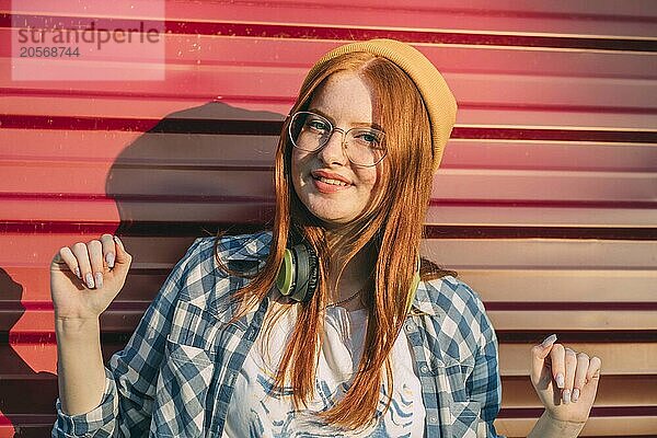 Red haired teenage girl in eyeglasses and wireless headphones near corrugated iron