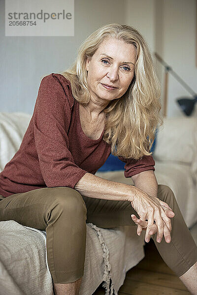 Smiling blond senior woman sitting on sofa in living room at home