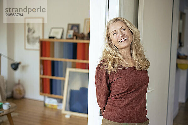 Smiling blond senior woman leaning on wall in living room at home