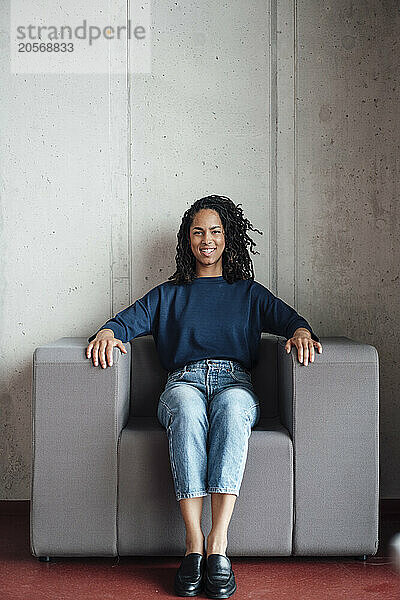 Smiling young businesswoman sitting on armchair in office lobby