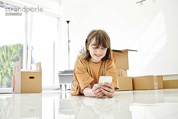 Smiling girl using smart phone lying on floor near boxes in new home