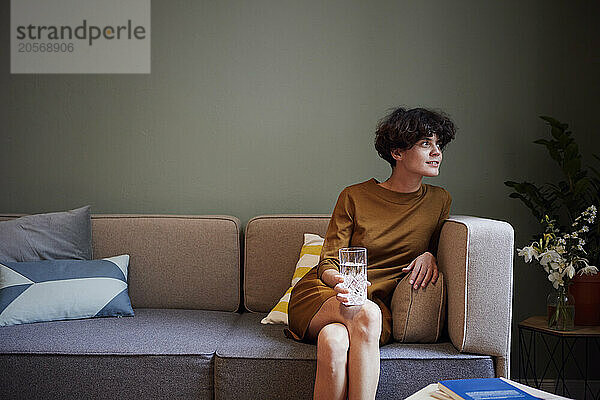 Thoughtful woman holding glass of water and sitting on sofa at home