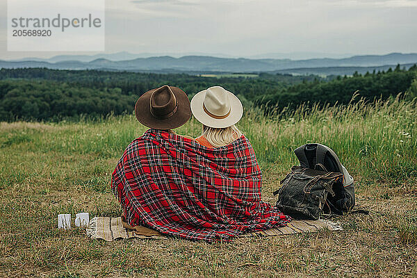 Young couple wearing hat and plaid shawl sitting on meadow at mountain