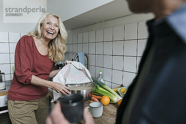 Cheerful senior woman showing mixing bowl to man standing in kitchen at home