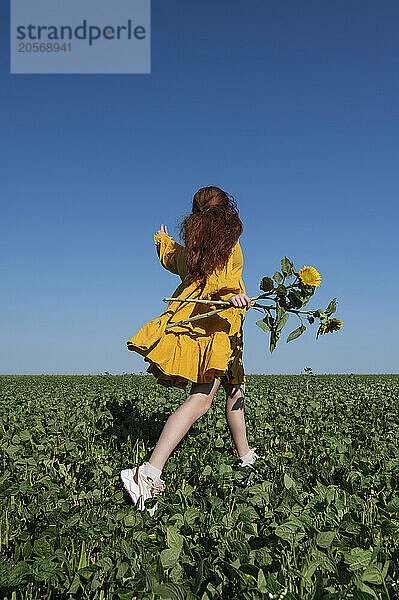 Teenage girl with red hair in a yellow dress running with sunflowers in a green field