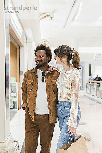 Happy multiracial couple with paper bag doing window shopping at mall