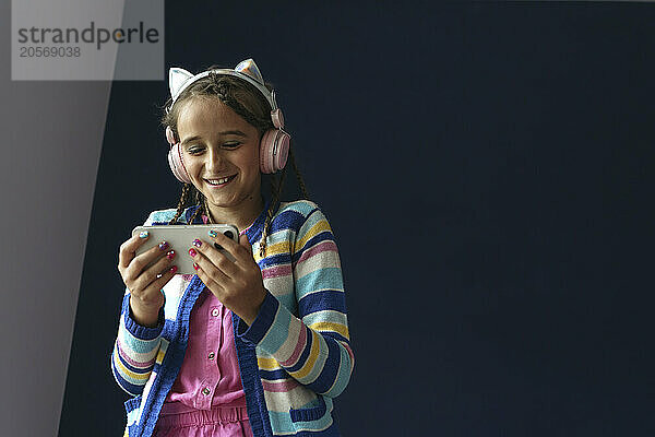 Happy girl listening music through headphones and using smart phone in front of wall at home