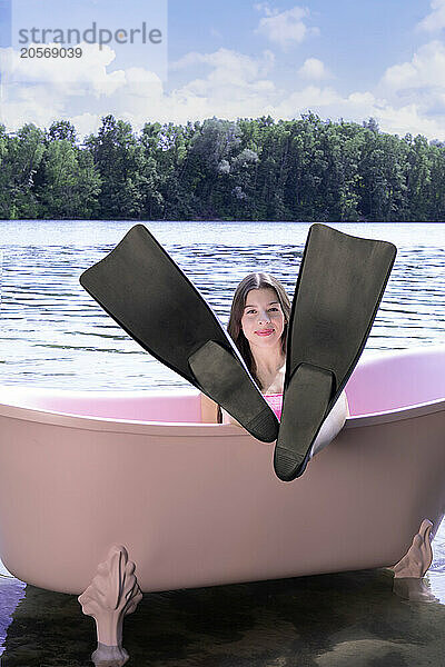 Smiling girl wearing diving flipper and sitting inside pink bathtub in lake