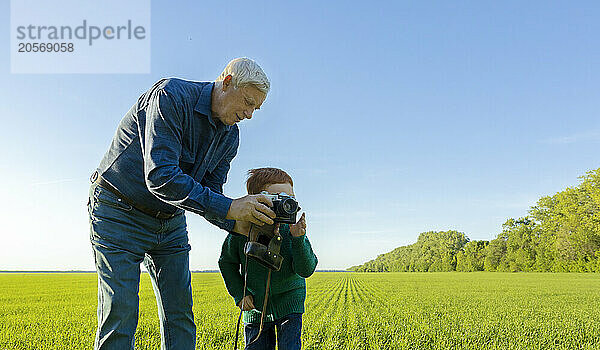 Boy looks into old camera by grandfather standing in field