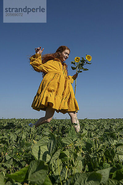 Teenage girl with red hair in a yellow dress running with sunflowers in a green field