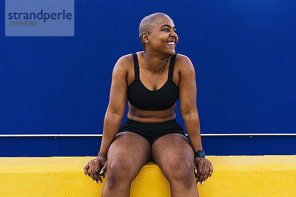 Smiling bald and muscular woman sitting in front of blue wall