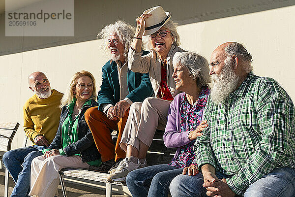 Cheerful woman wearing hat sitting amidst friends on bench in front of wall