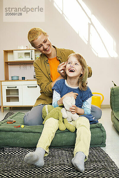 Smiling woman combing hair of daughter sitting on couch at home