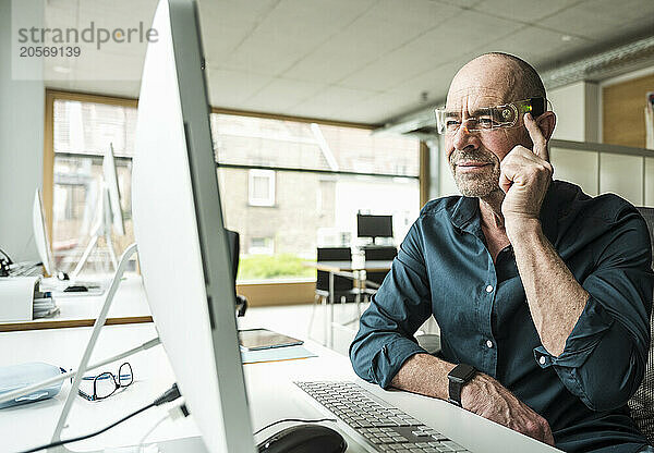 Confident mature businessman wearing smart glasses looking at computer monitor in office