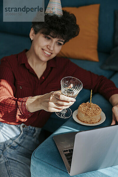 Smiling woman toasting wine and celebrating birthday through laptop at home