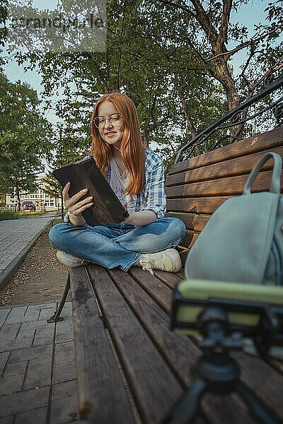 Smiling teenage girl using tablet PC sitting cross-legged on bench and vlogging through smart phone at park