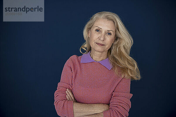 Confident blond senior woman with arms crossed in front of navy blue wall