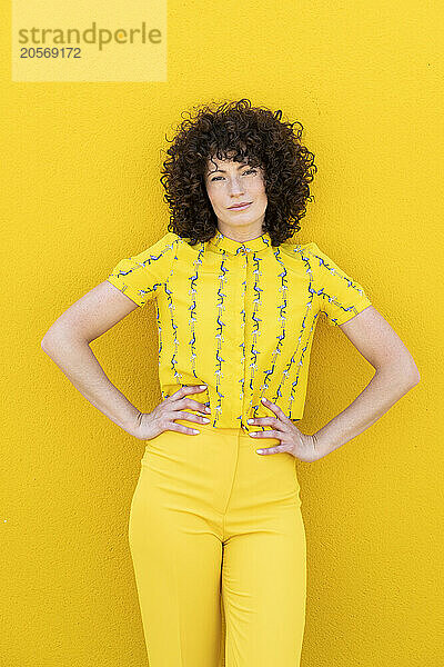 Confident woman standing with arms akimbo in front of yellow wall