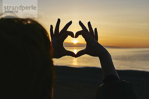 Woman making heart shape at beach during sunrise