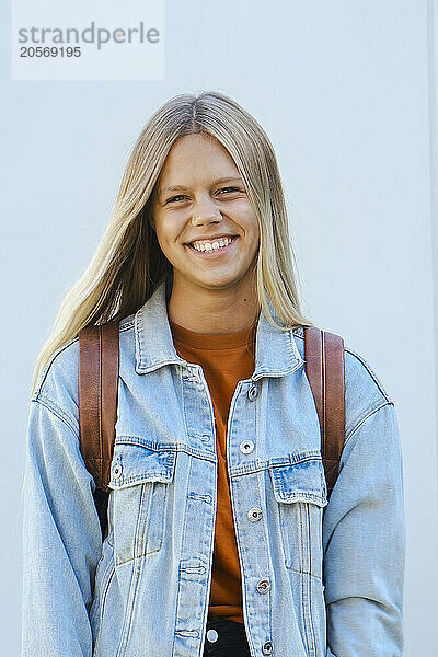 Happy blond hair teenager in denim jacket standing in front of wall