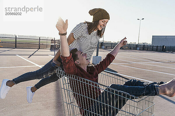 Playful woman pushing boyfriend sitting in shopping cart at parking lot