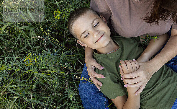 The boy lies on his mother's lap on the grass and she holds his hand.