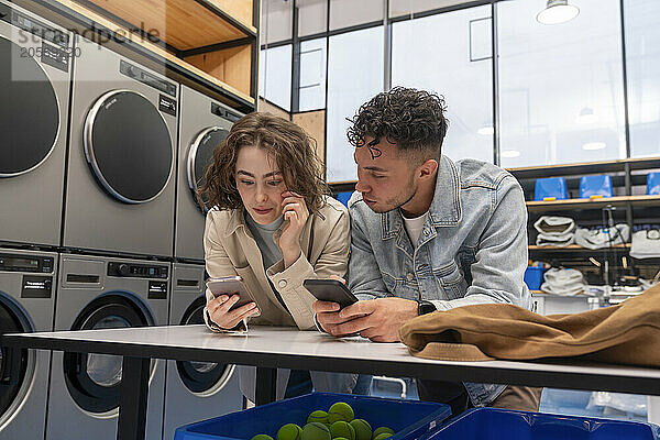 Young woman and man using smart phones leaning on table at laundromat