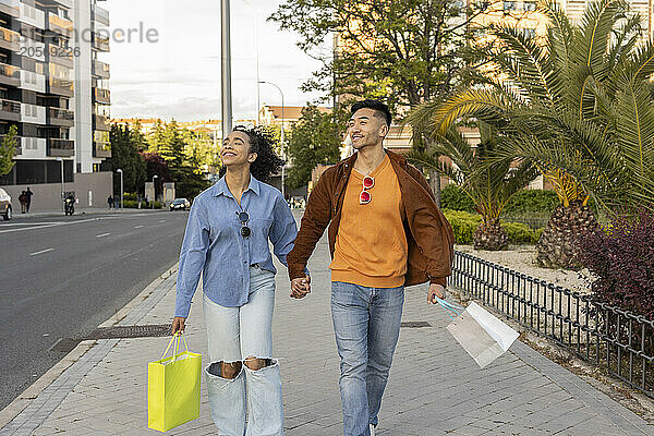 Happy multiracial couple carrying shopping bags and walking on footpath