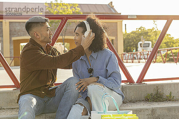 Young man helping girlfriend with wireless headphones and sitting on staircase at park