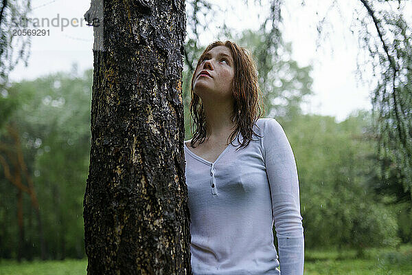 Woman looking up and standing near tree trunk in forest