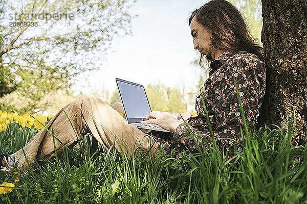 Man using laptop leaning on tree trunk at meadow