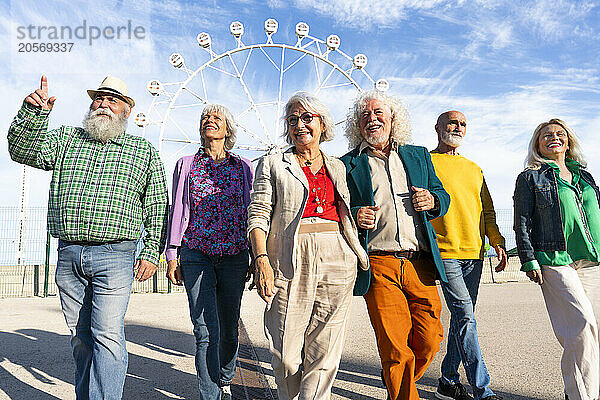 Retired men and women talking and walking side by side in front of Ferris wheel on sunny day