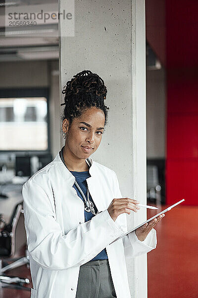 Confident young female doctor with tablet PC leaning on column in hospital lobby