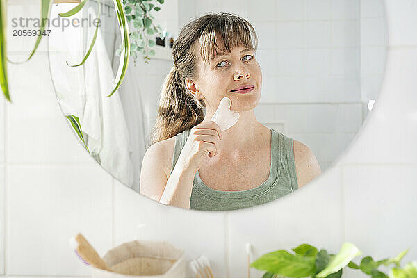 Beautiful mature woman massaging face with gua sha stone in bathroom