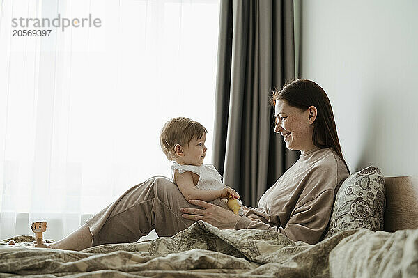 Smiling woman with daughter sitting on bed at home