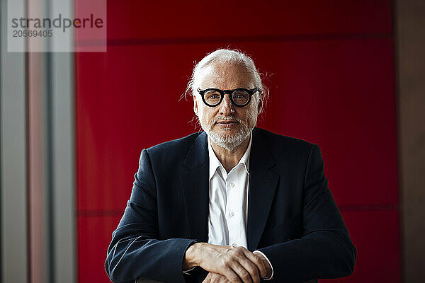 Confident senior businessman wearing eyeglasses in front of red wall at office