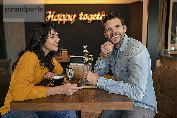 Happy businessman sitting with businesswoman holding mobile phone at table in cafe