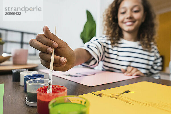 Girl dipping brush in paint pot sitting at table drawing