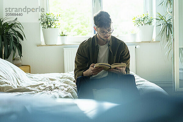 Young man reading book sitting on bed at home