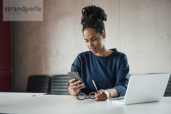 Young businesswoman with eyeglasses and laptop using smart phone at desk in office