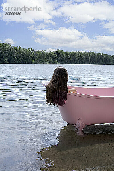 Girl with long hair sitting inside pink bathtub in lake