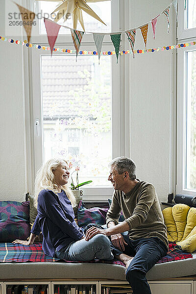 Smiling retired senior couple sitting on alcove window seat at home