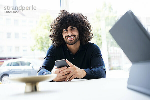 Smiling young curly haired businessman sitting with smart phone at desk in office