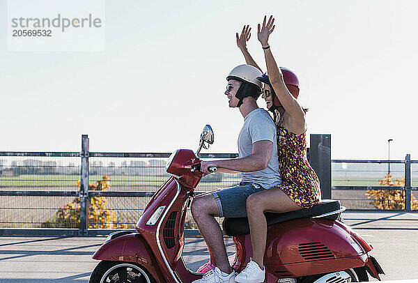 Cheerful young woman with arms raised sitting on motor scooter with boyfriend at parking lot