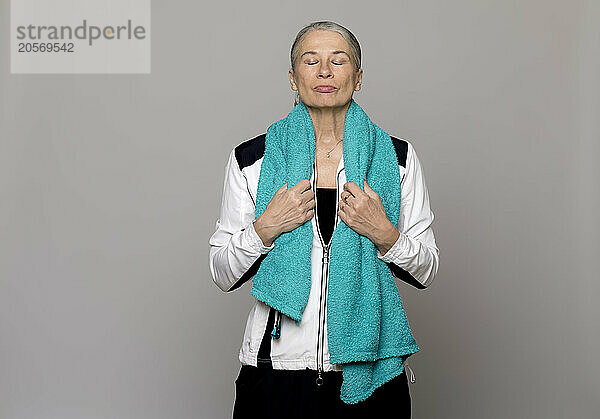 Senior woman with eyes closed and turquoise towel standing against gray background